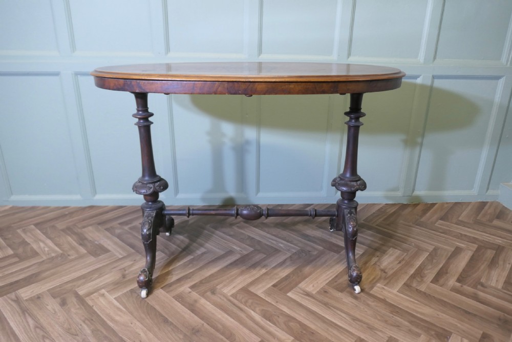 A Victorian oval walnut centre table featuring a polished timber top, turned support columns, a central stretcher, and carved legs with white ceramic castors.