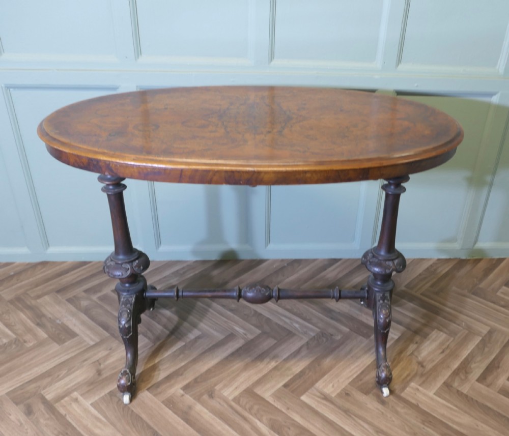 A Victorian oval walnut centre table featuring a polished timber top, turned support columns, a central stretcher, and carved legs with white ceramic castors. View II