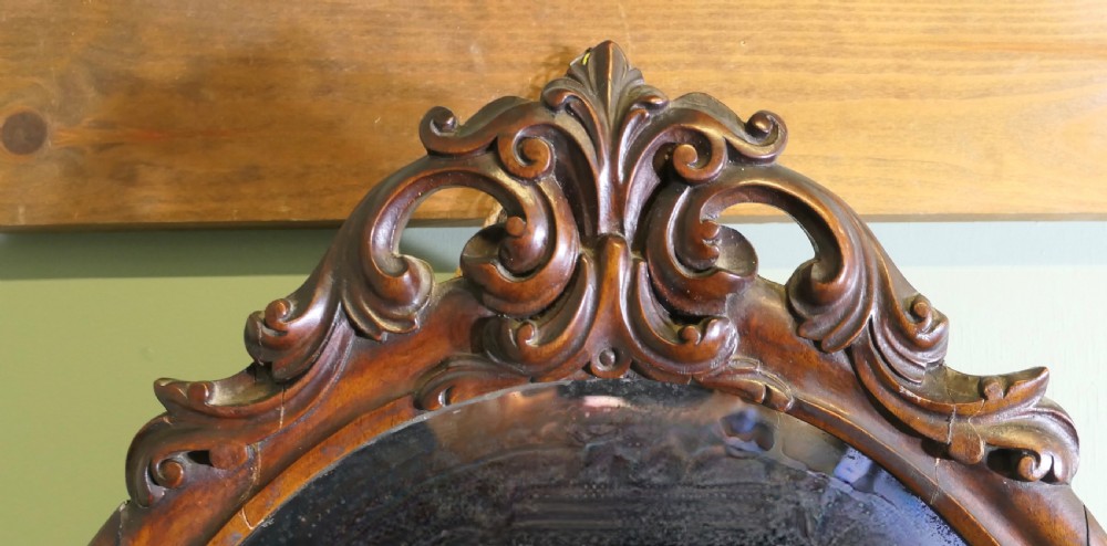 An antique oval wall mirror featuring a moulded dark timber frame topped with an ornate carved scrollwork pediment. View II