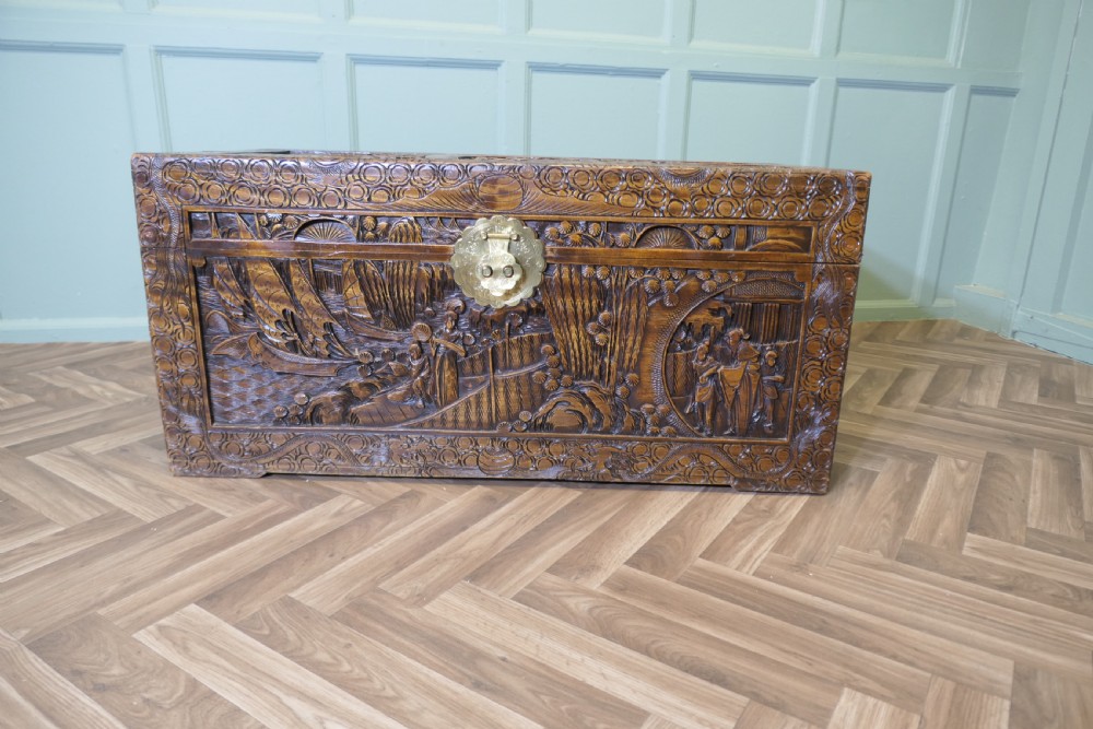 A large oriental camphor wood chest with detailed relief carvings depicting figures and sailing ships, complete with a central brass latch.