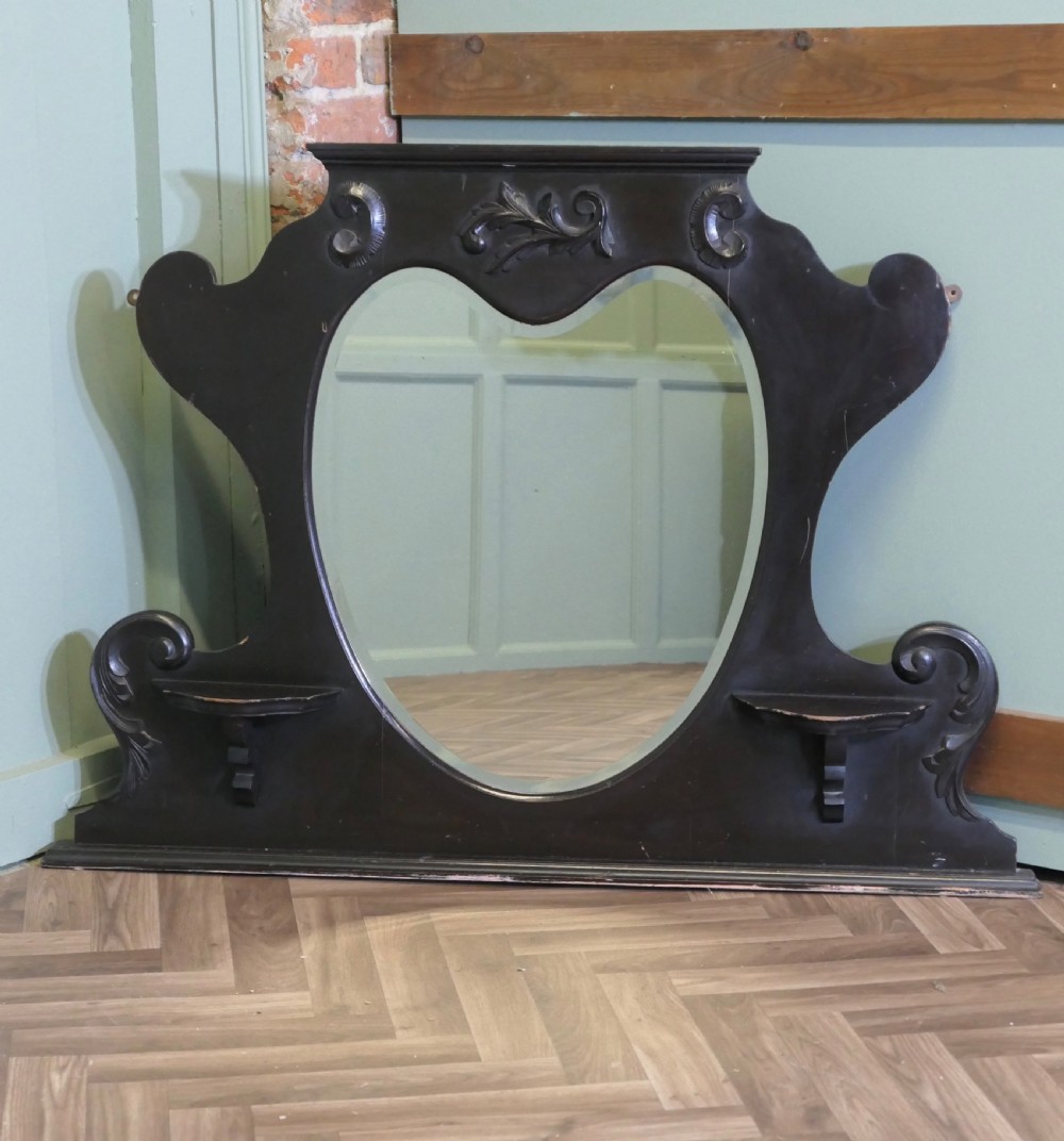 An Edwardian black lacquered overmantel mirror featuring a central heart-shaped glass plate, carved leaf crest, and two integrated side display shelves.