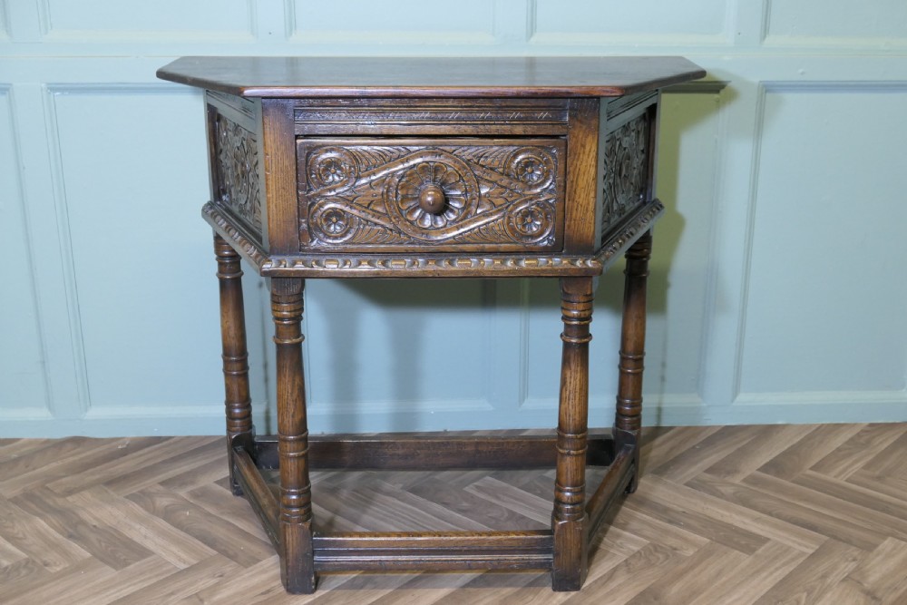 A dark oak credence table featuring a canted top, a single relief-carved drawer, turned legs, and a lower stretcher base.