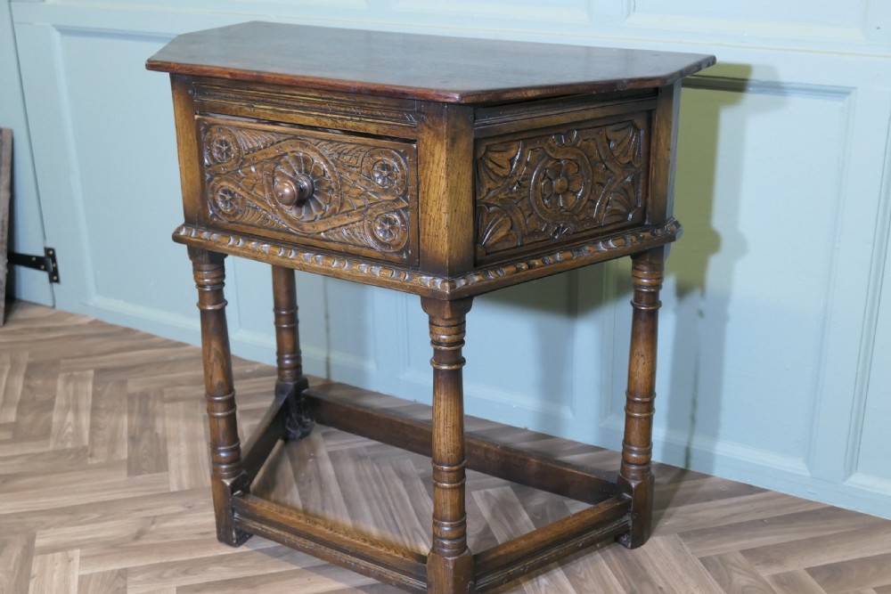 A dark oak credence table featuring a canted top, a single relief-carved drawer, turned legs, and a lower stretcher base. View III