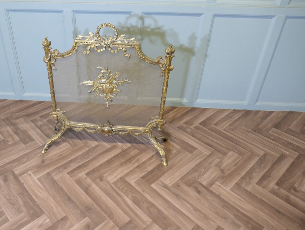 An ornate French gilded brass fire screen featuring intricate floral swags, a central bow motif, fine mesh panel, and decorative cabriole feet.