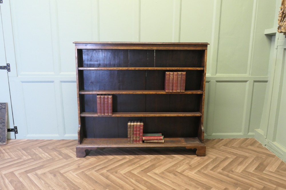 A wide antique dark elm open bookcase featuring four horizontal shelves, a panelled back, and bracket feet.