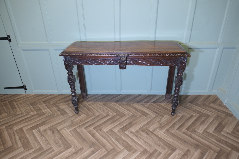 A dark carved oak hall table featuring an intricately carved apron with a central mask motif and turned front legs.