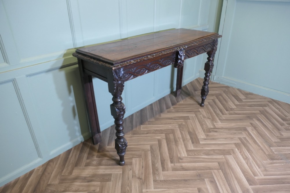 A dark carved oak hall table featuring an intricately carved apron with a central mask motif and turned front legs. View V