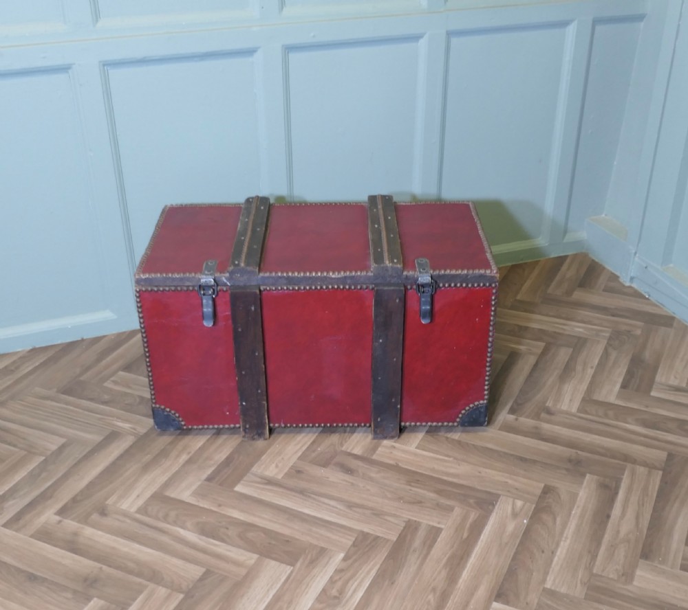 A rectangular dark red leather travel trunk with brass stud trimming, heavy dark wood banding, and iron metal latches resting on a parquet floor.