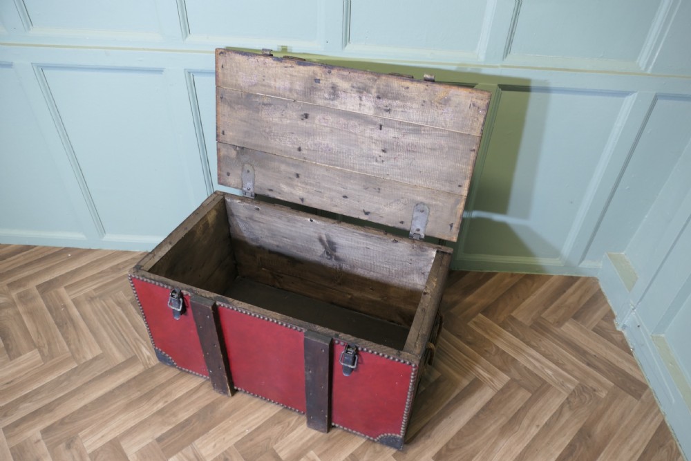 A rectangular dark red leather travel trunk with brass stud trimming, heavy dark wood banding, and iron metal latches resting on a parquet floor. View V