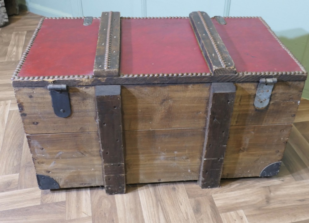 A rectangular dark red leather travel trunk with brass stud trimming, heavy dark wood banding, and iron metal latches resting on a parquet floor. View VI