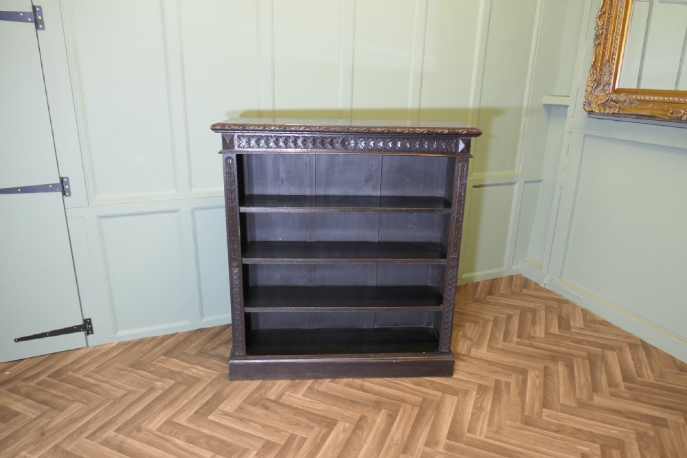 A dark carved oak open bookcase featuring four exposed shelves, a relief carved top frieze, and carved vertical side pilasters.