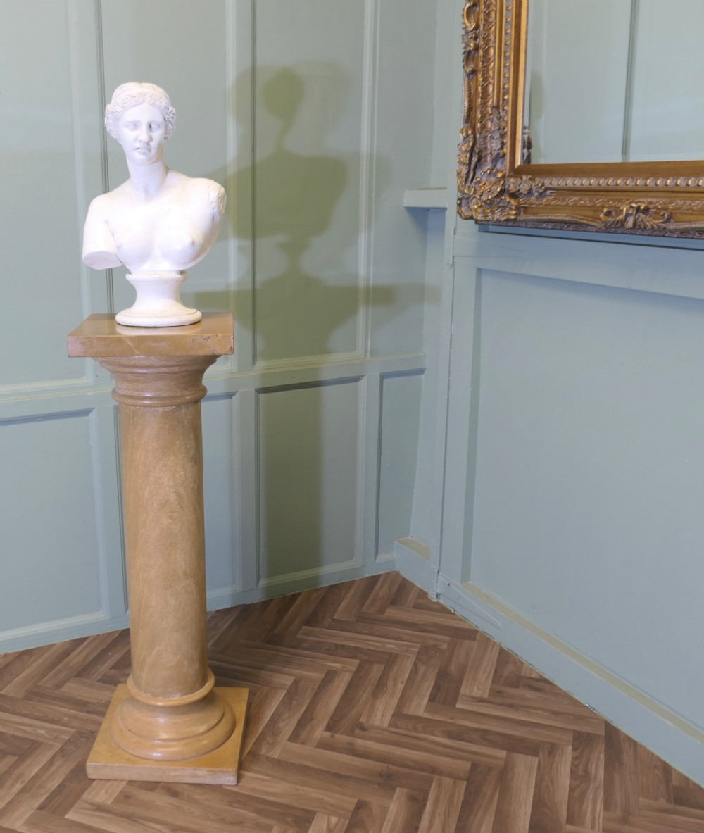 A tall honey-coloured marble column pedestal with a square top platform, cylindrical turned shaft, and square plinth base. View V
