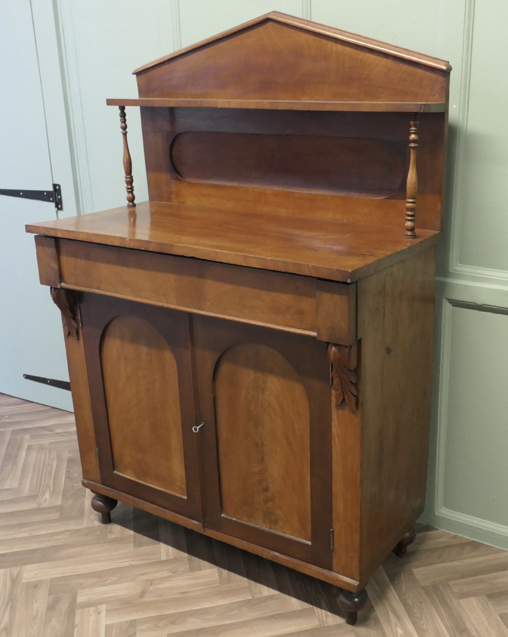 A Victorian mahogany chiffonier featuring a pedimented backboard with a shelf, turned supports, twin arched panel doors, and bun feet.