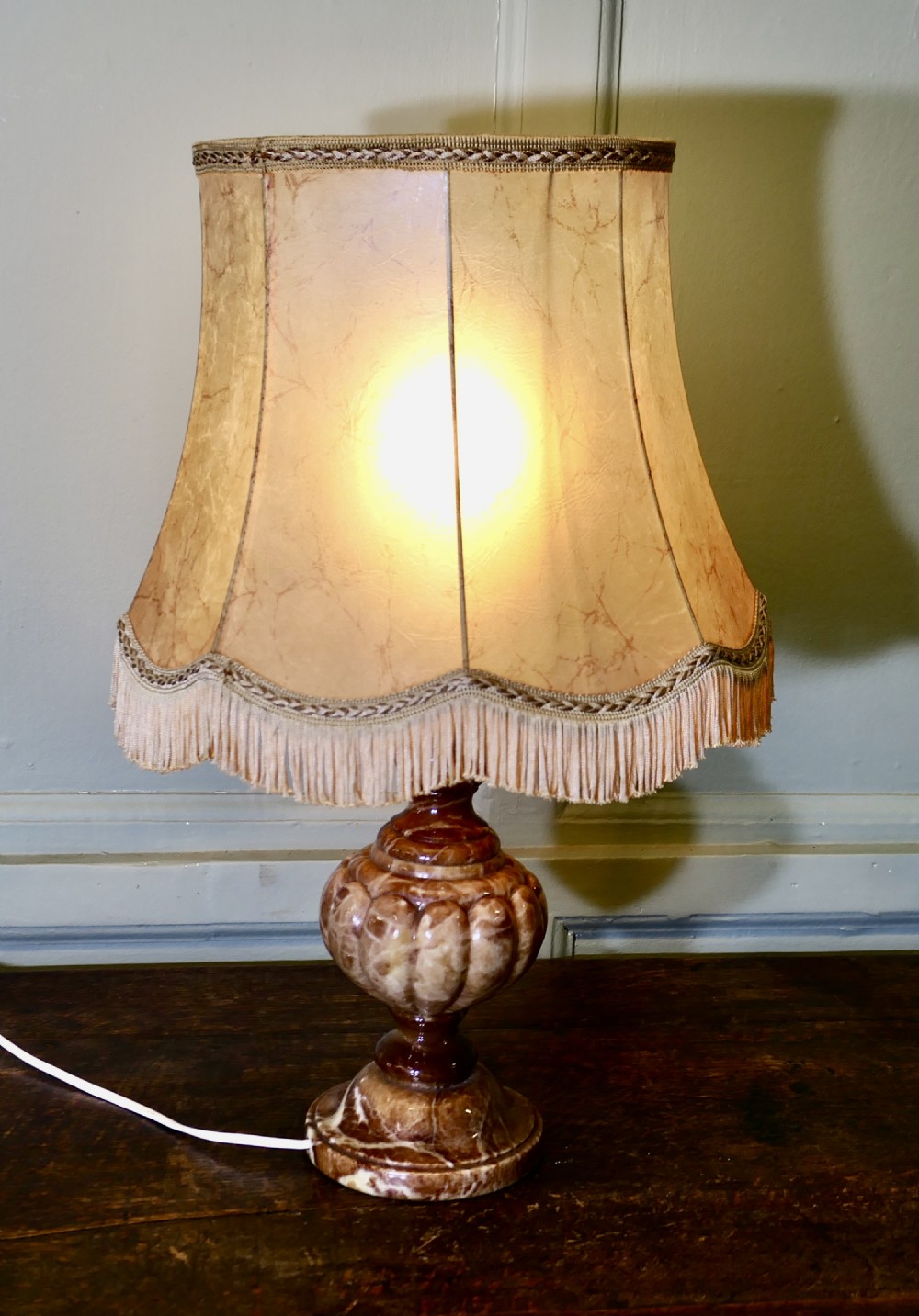 An Art Deco table lamp featuring a carved variegated brown marble urn base and a flared beige lampshade with fringe details.