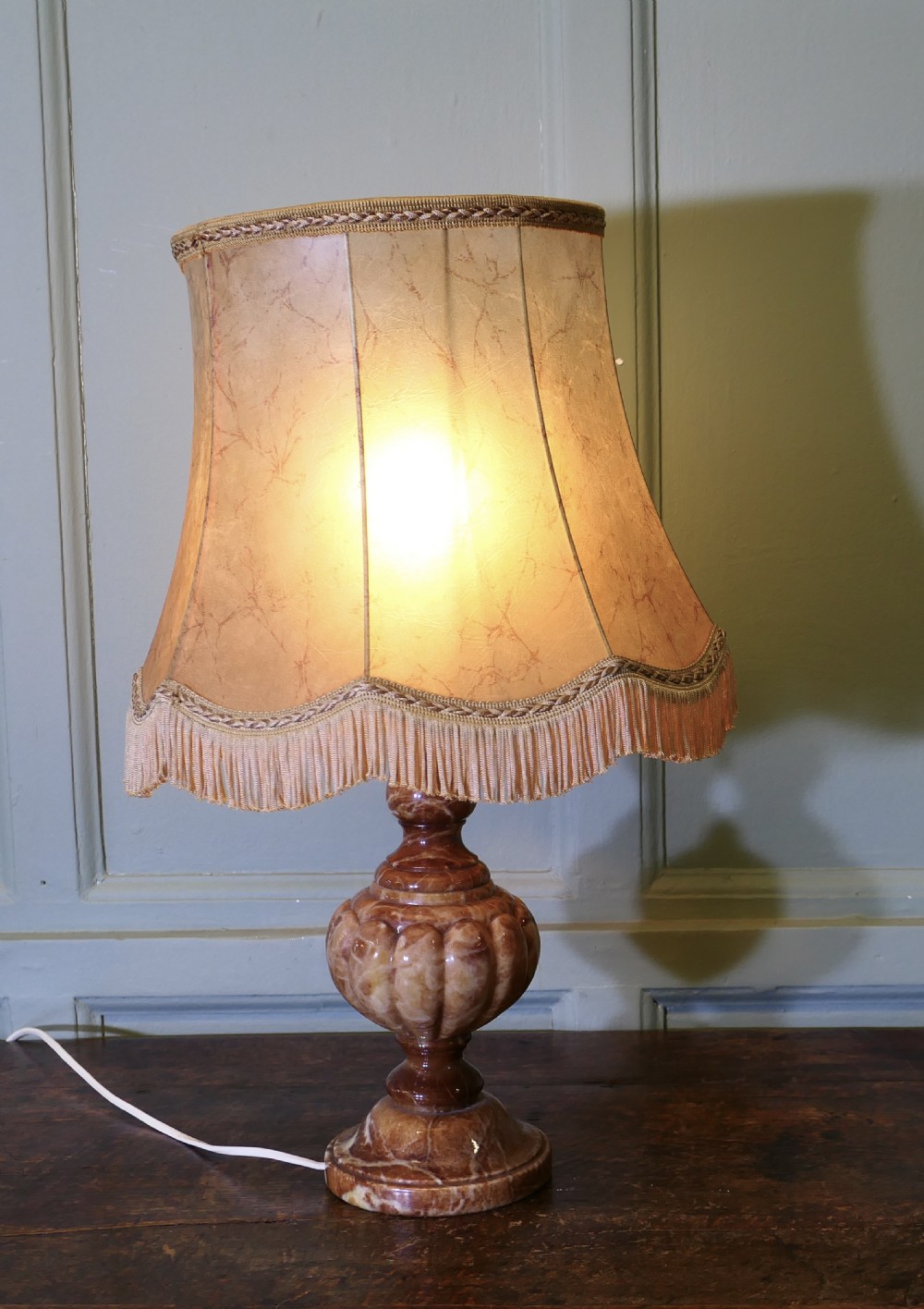 An Art Deco table lamp featuring a carved variegated brown marble urn base and a flared beige lampshade with fringe details. View V