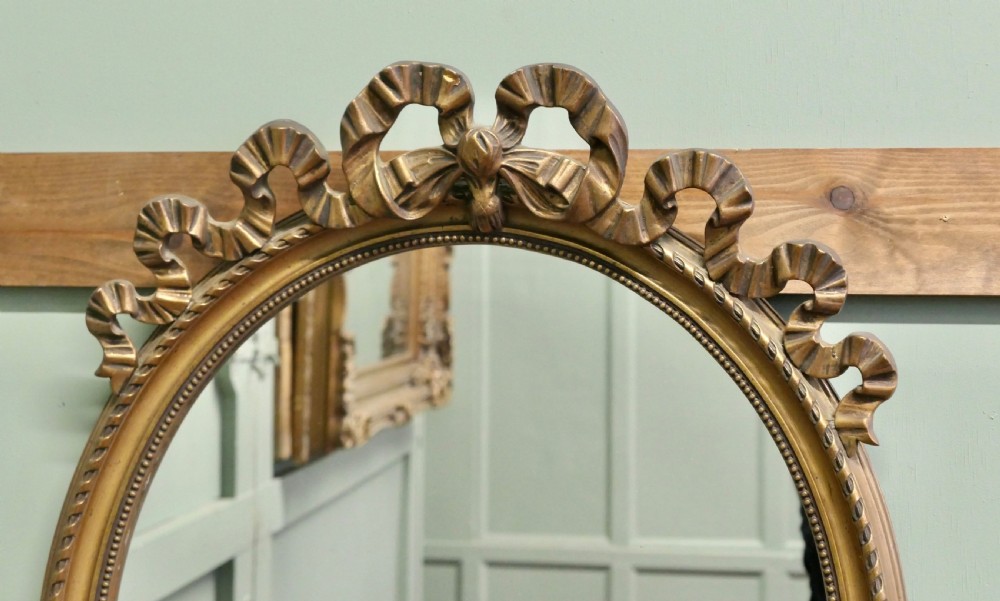 An oval wall mirror with a gilded frame topped with a bow ribbon motif. View III