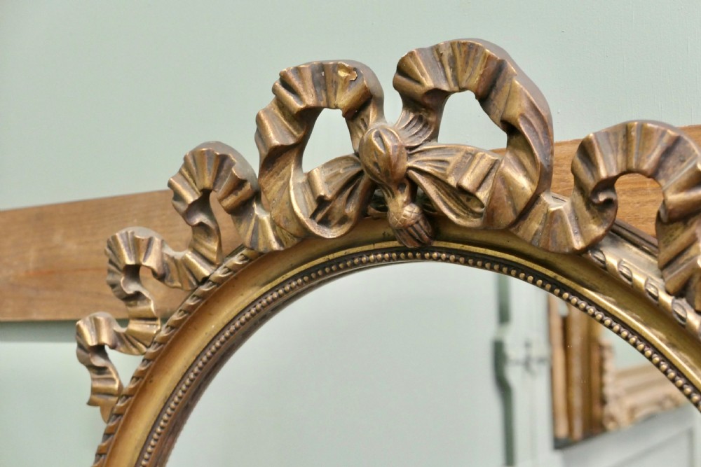 An oval wall mirror with a gilded frame topped with a bow ribbon motif. View IV