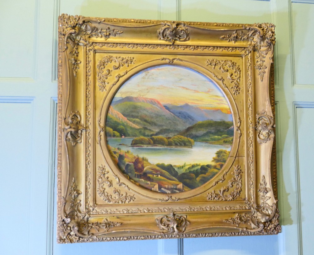A Victorian oil landscape painting depicting mountains and a lake, set inside a circular aperture framed by a heavy square gilded gesso frame.