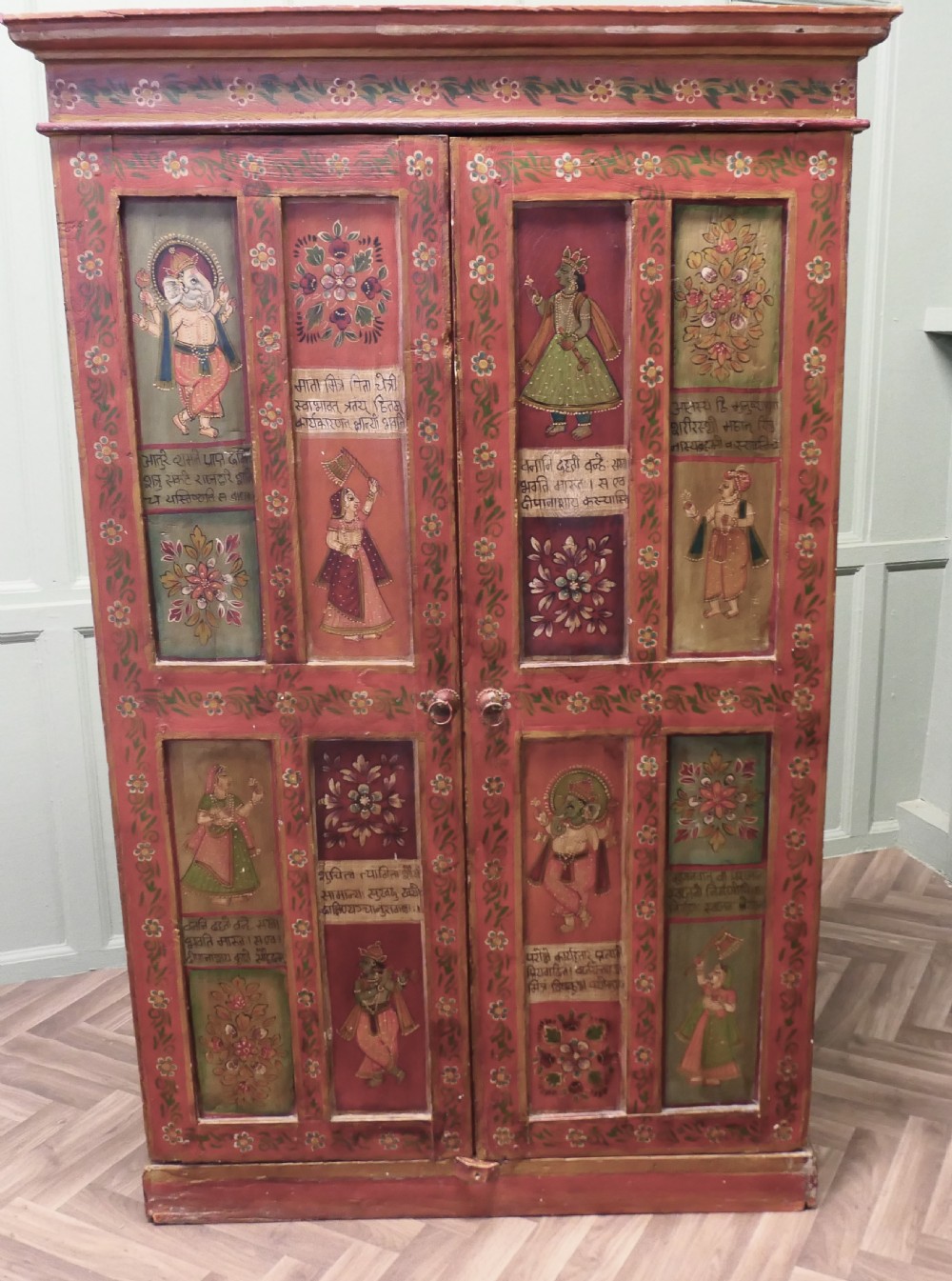 A tall red hand-painted Indian wooden cupboard decorated with framed panels featuring deities, figure artwork, Devanagari script, and floral motifs.