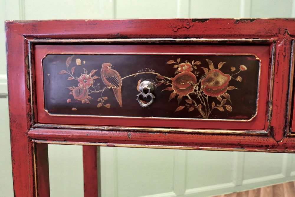A red lacquered rectangular console table featuring two dark drawers decorated with painted floral motifs and metal ring handles on square legs. View III