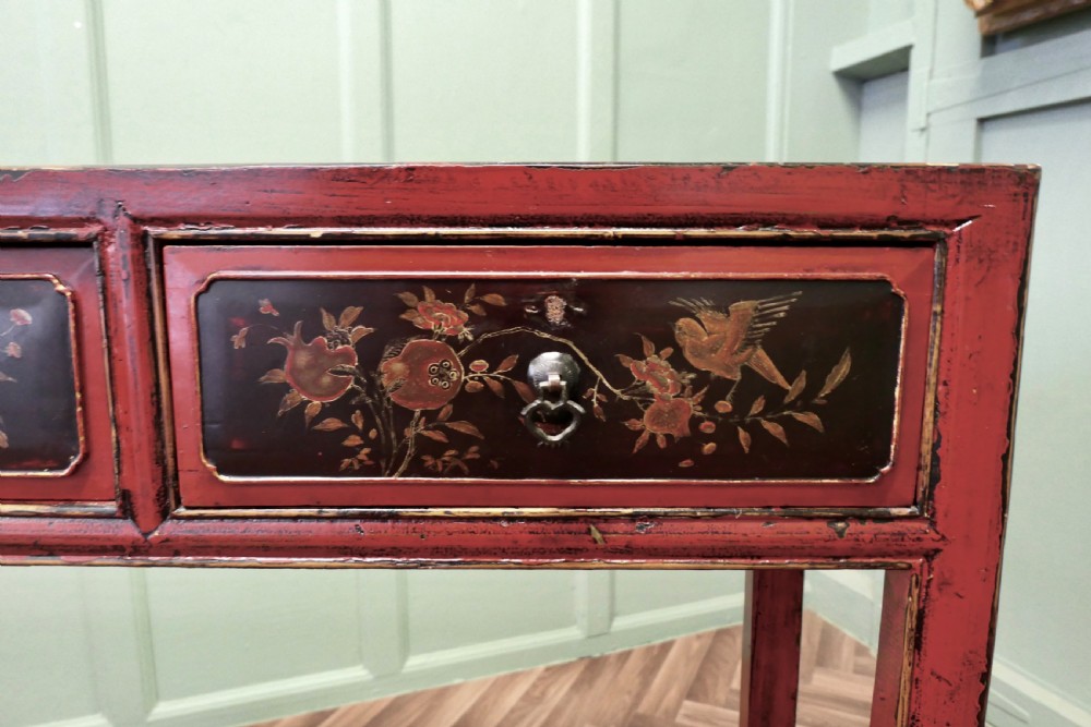 A red lacquered rectangular console table featuring two dark drawers decorated with painted floral motifs and metal ring handles on square legs. View V