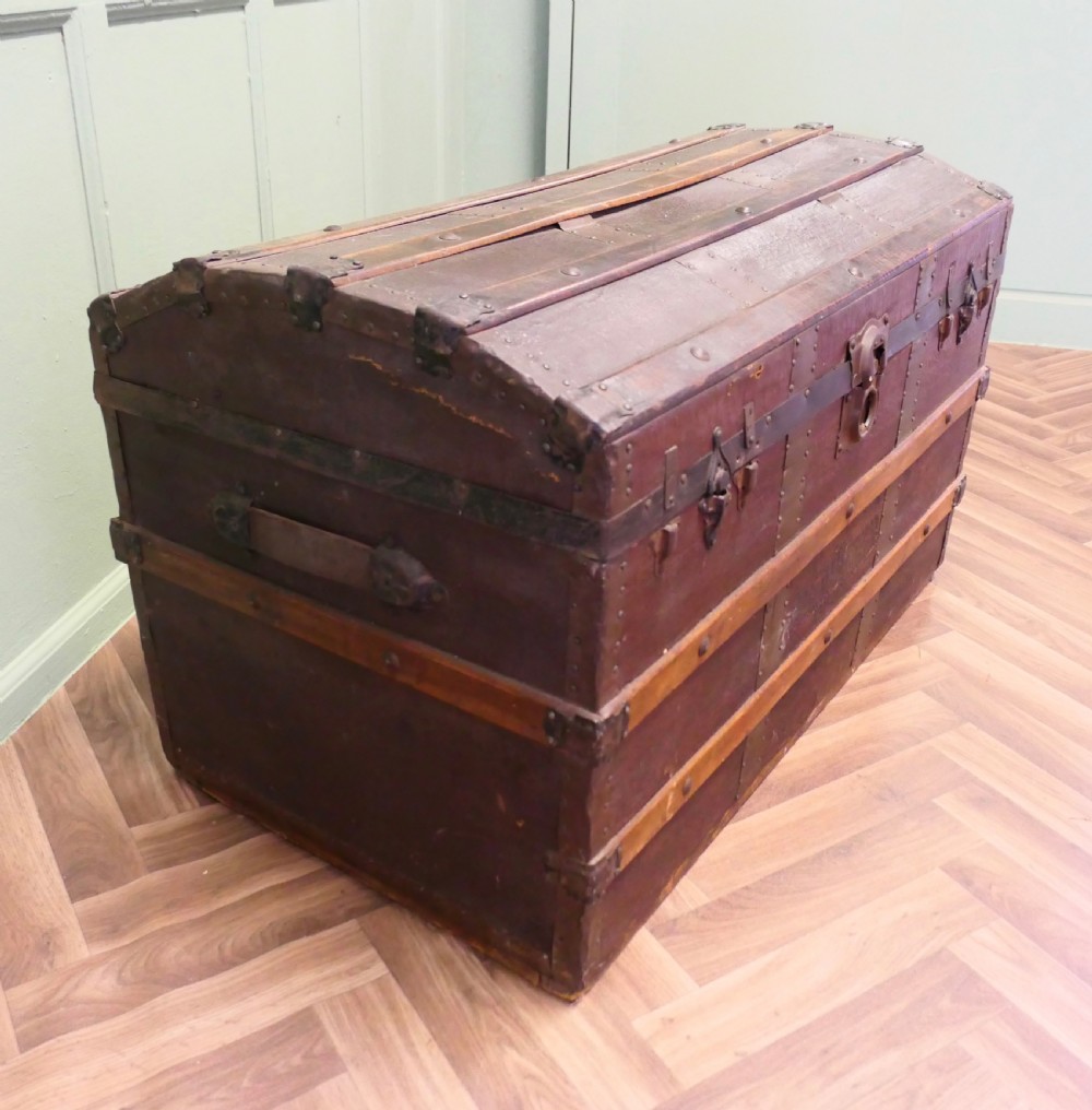 An antique dome top steamer trunk featuring dark canvas casing, polished oak slats, metal banding, and side leather handles.
