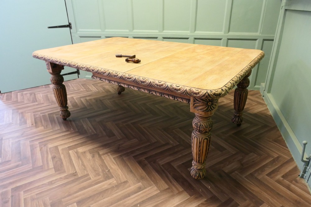 A rectangular bleached oak extending dining table with ornate carved aprons and turned legs.