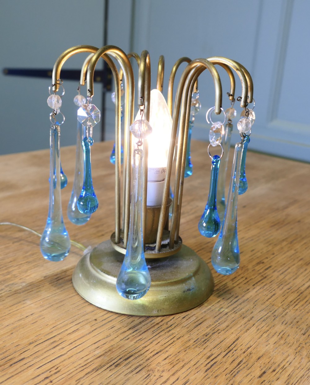 A small brass table lamp featuring curved fountain-like arms adorned with hanging turquoise and clear glass teardrop droplets surrounding a central bulb. View III
