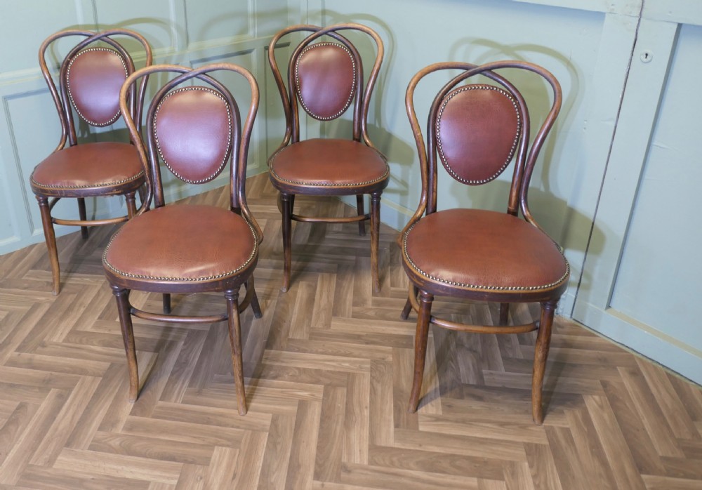 A set of four bentwood bistro dining chairs featuring steam-bent wooden frames and brown leather upholstery detailed with brass studding.
