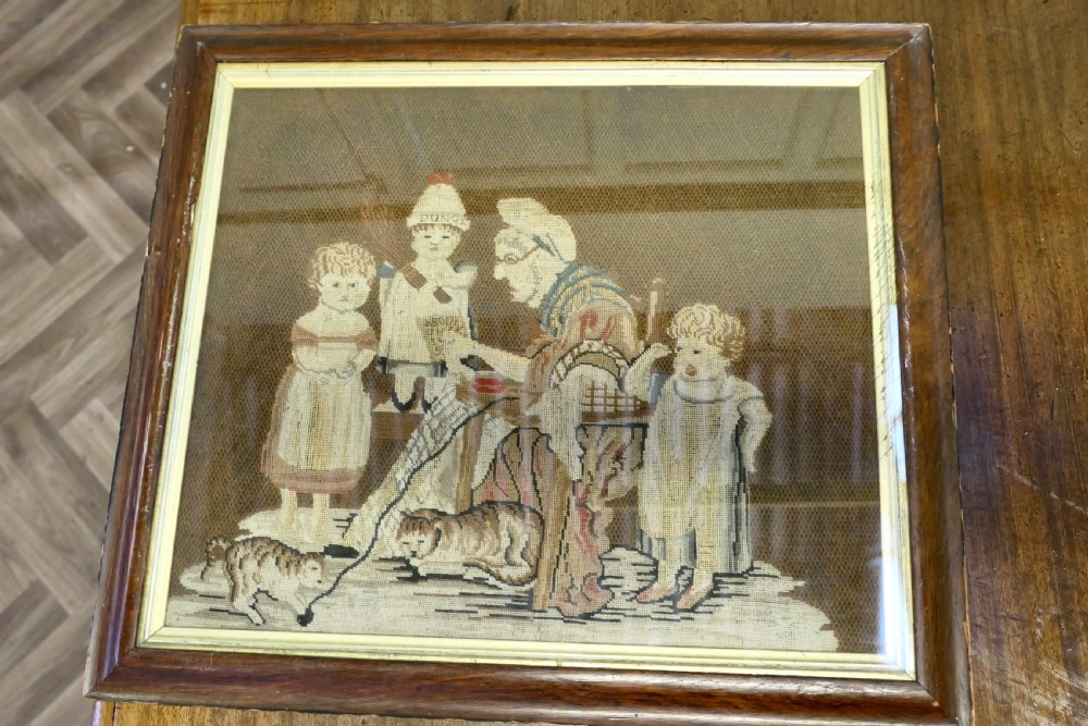 A framed early nineteenth-century wool needlework depicting a classroom scene with a seated teacher, children, and cats.