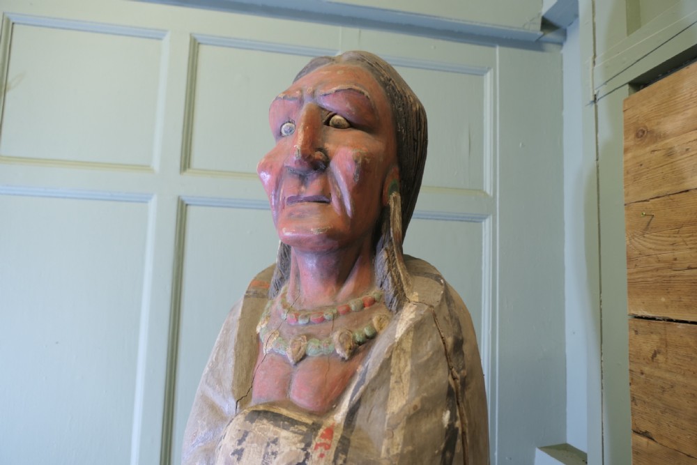 A tall, hand-carved painted wooden figure of a Native American wearing a patterned blanket and bead necklace, standing on an integral round timber base. View VI