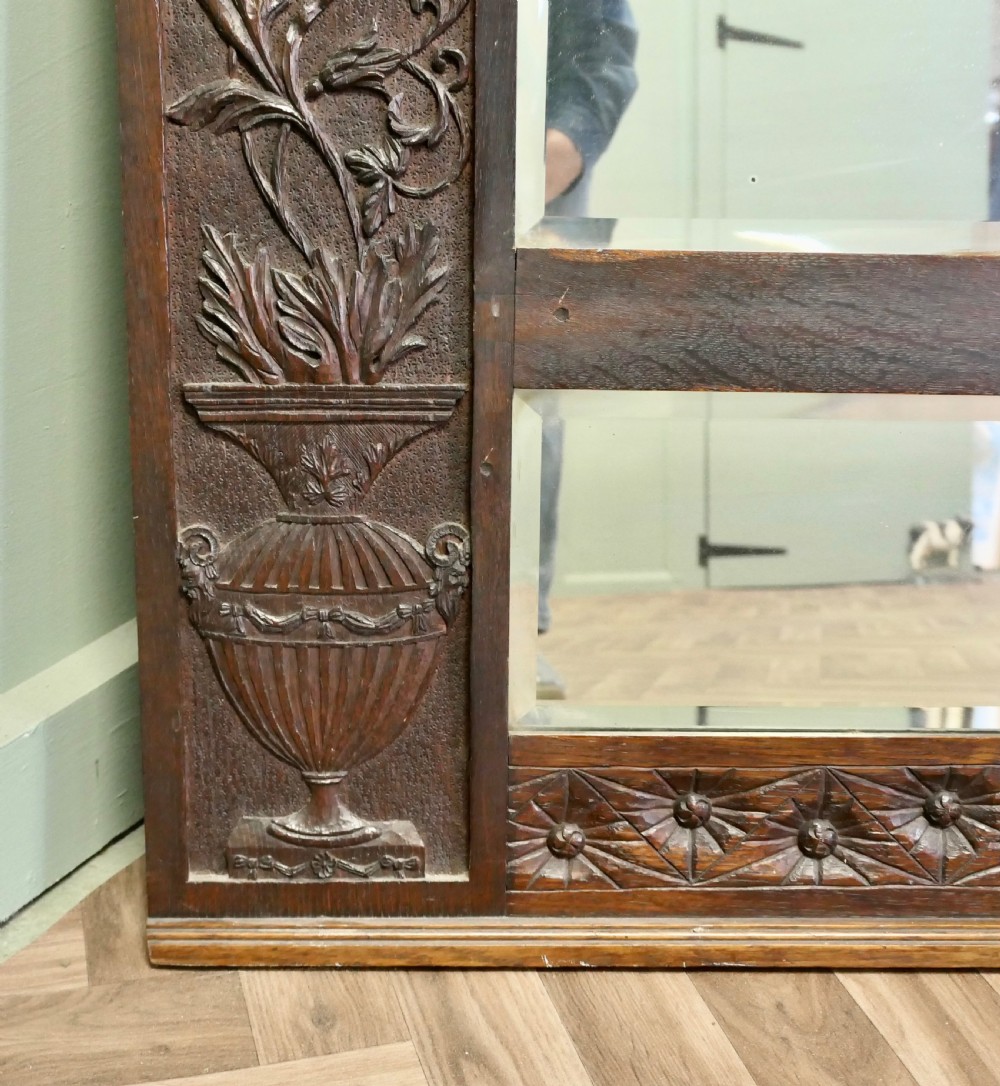 A large rectangular dark oak wall mirror featuring hand-carved foliate side panels, carved urn motifs, a rosette border, and two glass panes. View VI