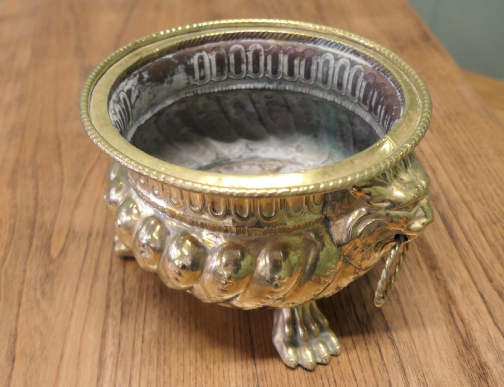 An ornate round brass jardiniere featuring gadrooned ribbed sides, lion mask ring handles, and paw feet.