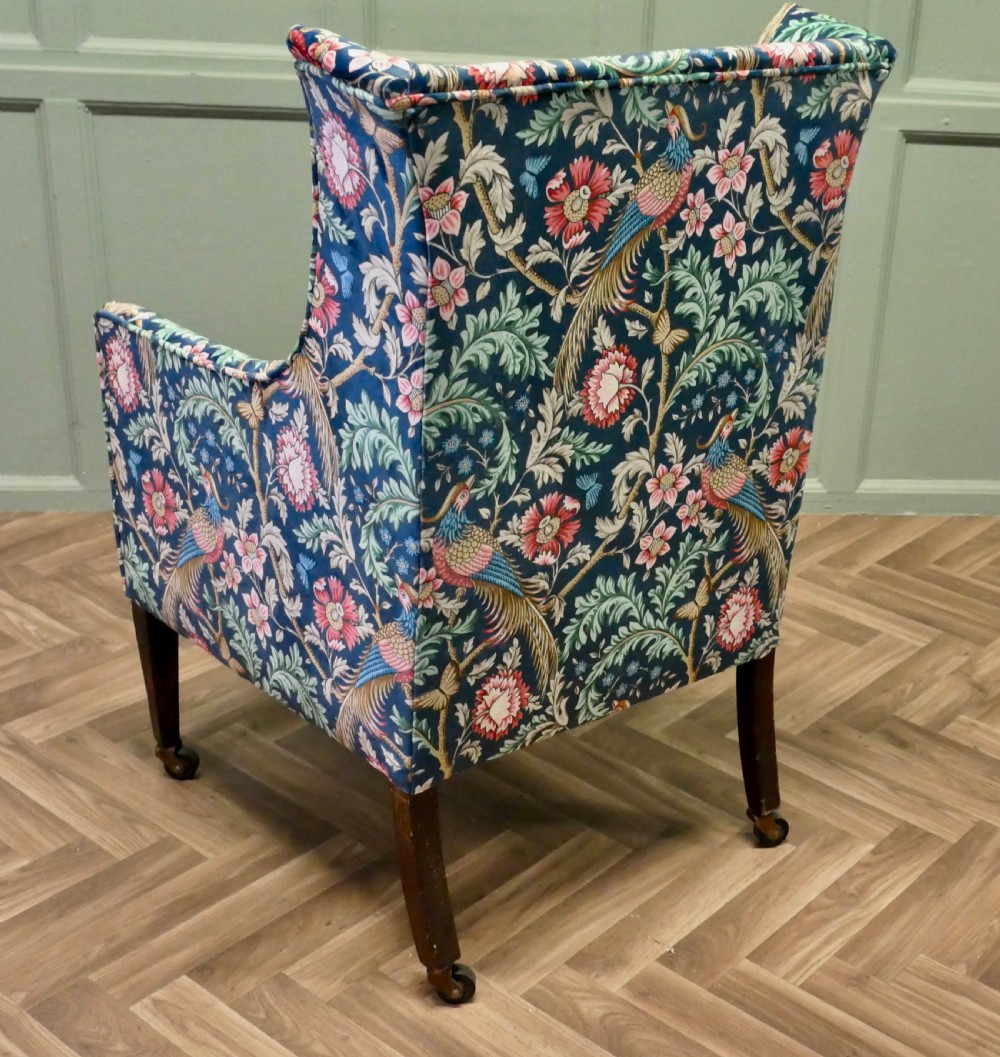 A high-backed wingback armchair upholstered in navy blue velvet featuring a colourful botanical and bird pattern, standing on dark timber legs with brass castors. View III