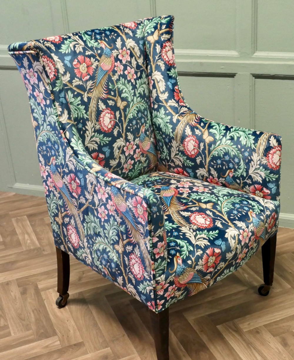 A high-backed wingback armchair upholstered in navy blue velvet featuring a colourful botanical and bird pattern, standing on dark timber legs with brass castors. View V