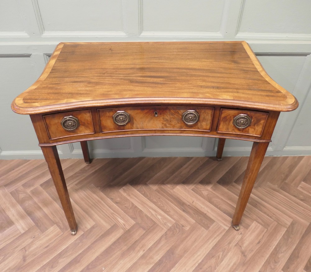 A Victorian walnut writing table featuring a concave front, crossbanded top surface, four brass rosette drawer handles, and square tapered legs on brass castors.