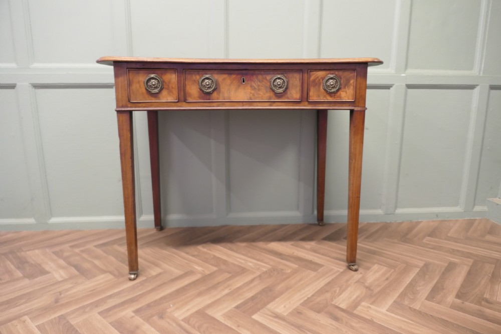 A Victorian walnut writing table featuring a concave front, crossbanded top surface, four brass rosette drawer handles, and square tapered legs on brass castors. View II