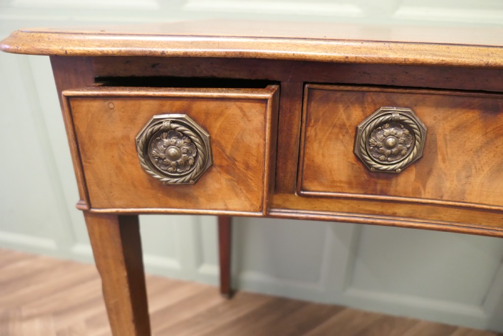 A Victorian walnut writing table featuring a concave front, crossbanded top surface, four brass rosette drawer handles, and square tapered legs on brass castors. View III
