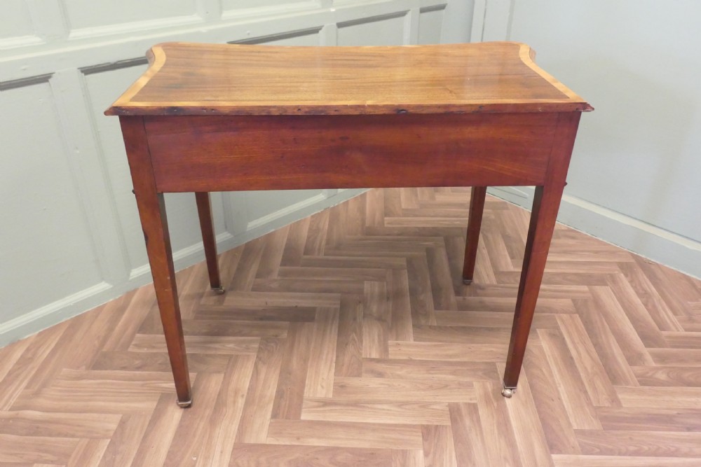A Victorian walnut writing table featuring a concave front, crossbanded top surface, four brass rosette drawer handles, and square tapered legs on brass castors. View V