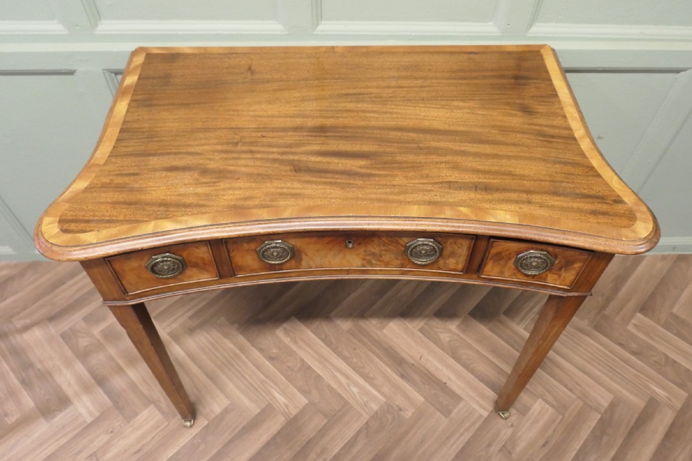 A Victorian walnut writing table featuring a concave front, crossbanded top surface, four brass rosette drawer handles, and square tapered legs on brass castors. View VI