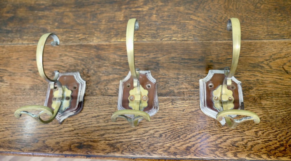 A set of three French artisan brass coat hooks featuring curved double arms mounted onto shield-shaped backplates.