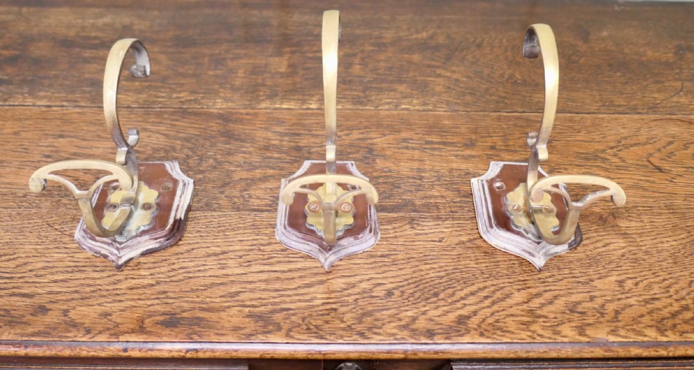 A set of three French artisan brass coat hooks featuring curved double arms mounted onto shield-shaped backplates. View II