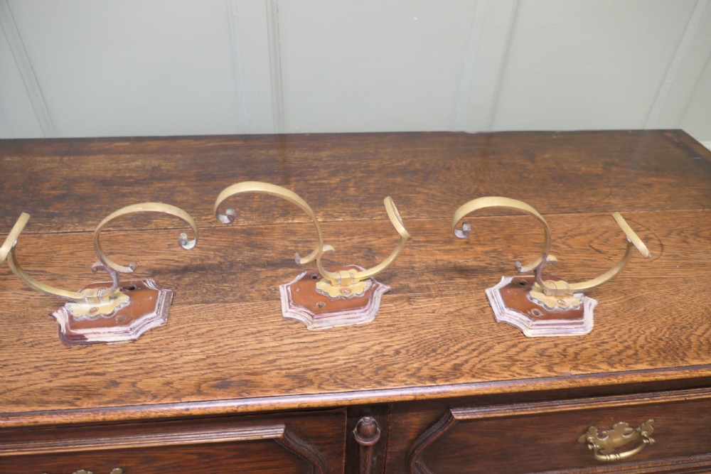 A set of three French artisan brass coat hooks featuring curved double arms mounted onto shield-shaped backplates. View V