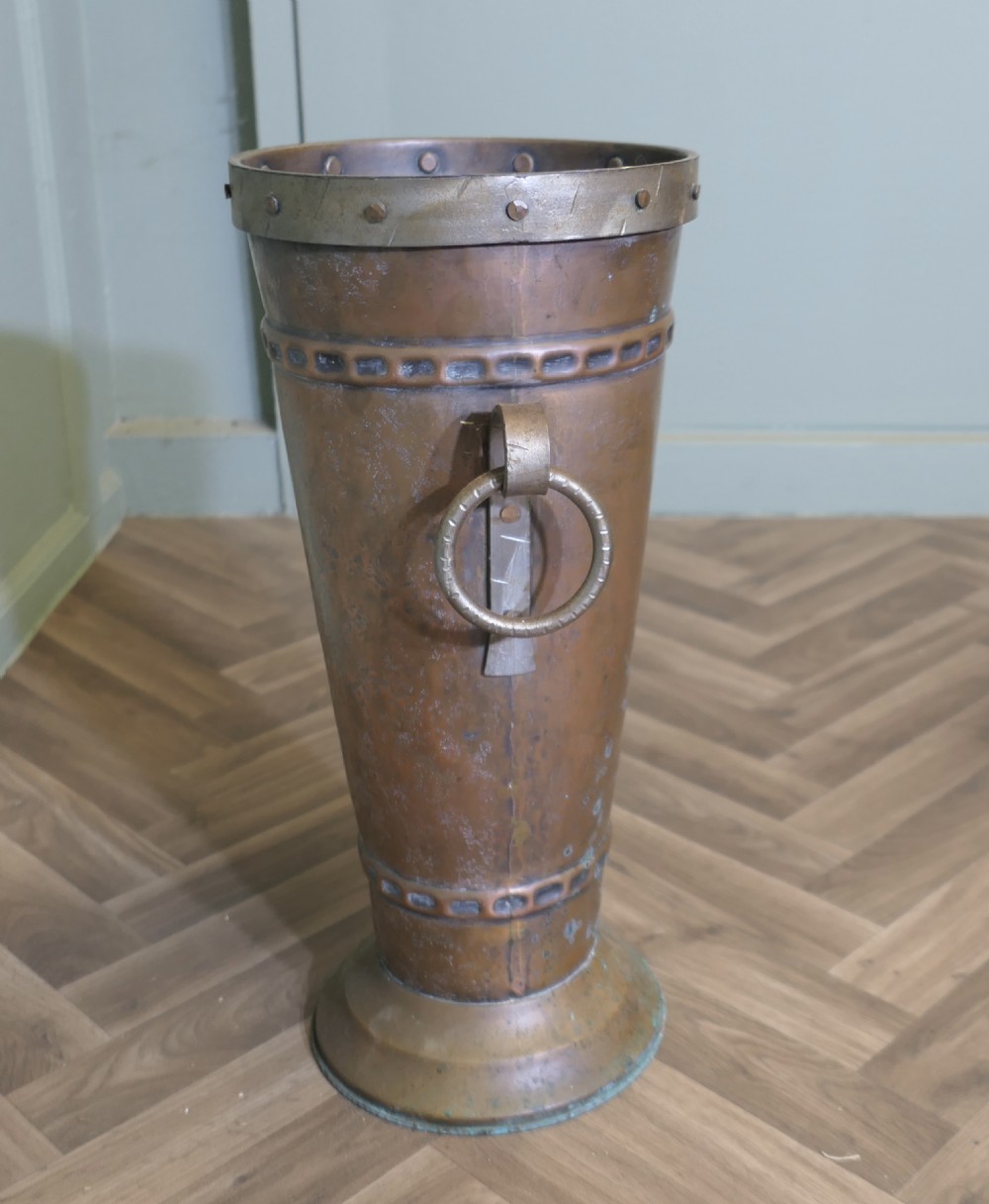 A tall hand-hammered copper umbrella stand featuring riveted metal trim, decorative embossed bands, and side ring handles on a flared circular base. View V