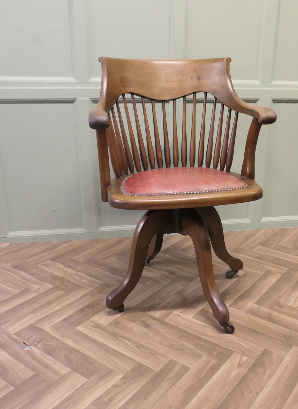 An Arts and Crafts wooden swivel desk chair with a spindle back, curved armrests, a studded leather seat, and a quadruped base with castors.