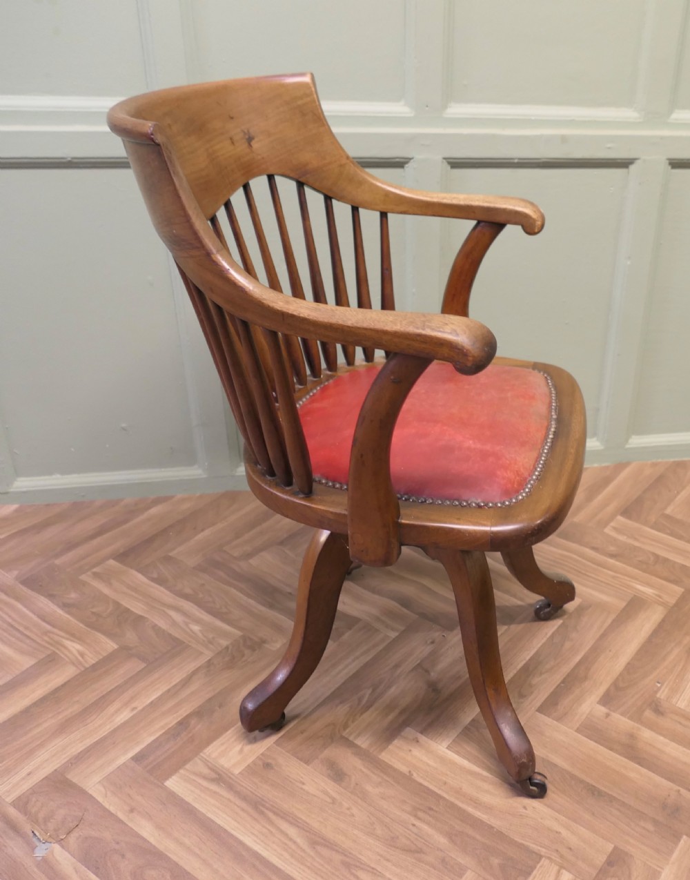 An Arts and Crafts wooden swivel desk chair with a spindle back, curved armrests, a studded leather seat, and a quadruped base with castors. View II