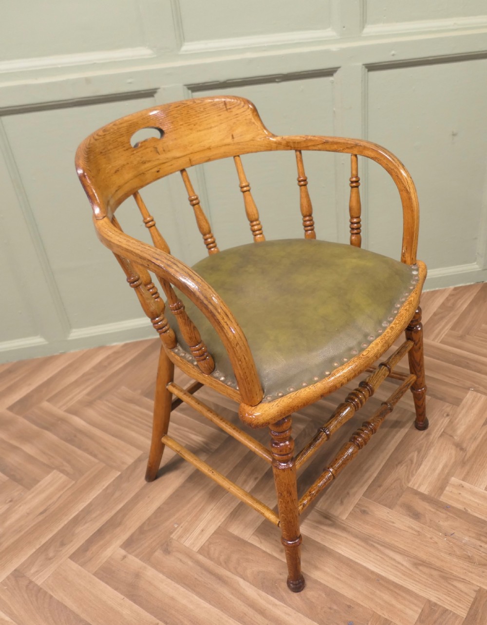 A golden oak smokers bow armchair featuring turned spindles, a steam-bent curved top rail, double stretchers, and a padded green leather seat secured with studs. View II