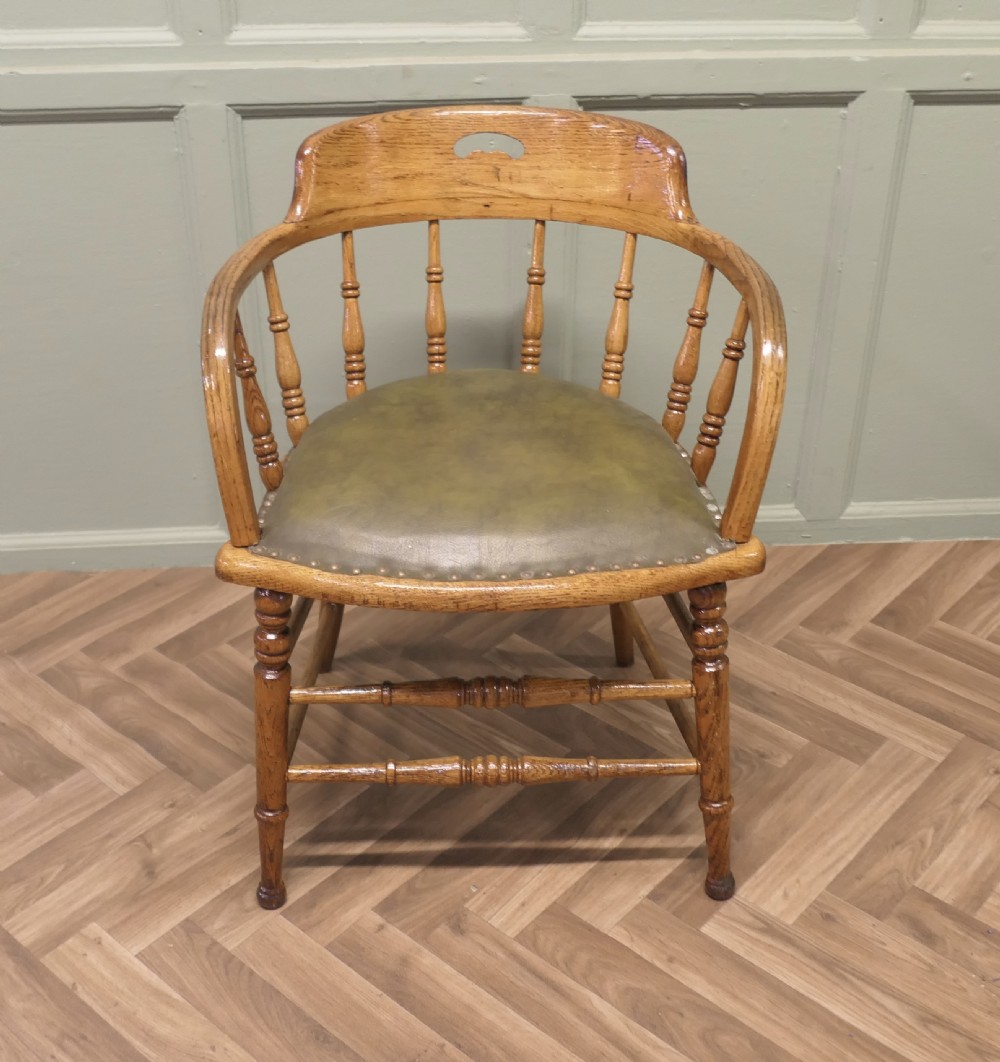 A golden oak smokers bow armchair featuring turned spindles, a steam-bent curved top rail, double stretchers, and a padded green leather seat secured with studs. View III