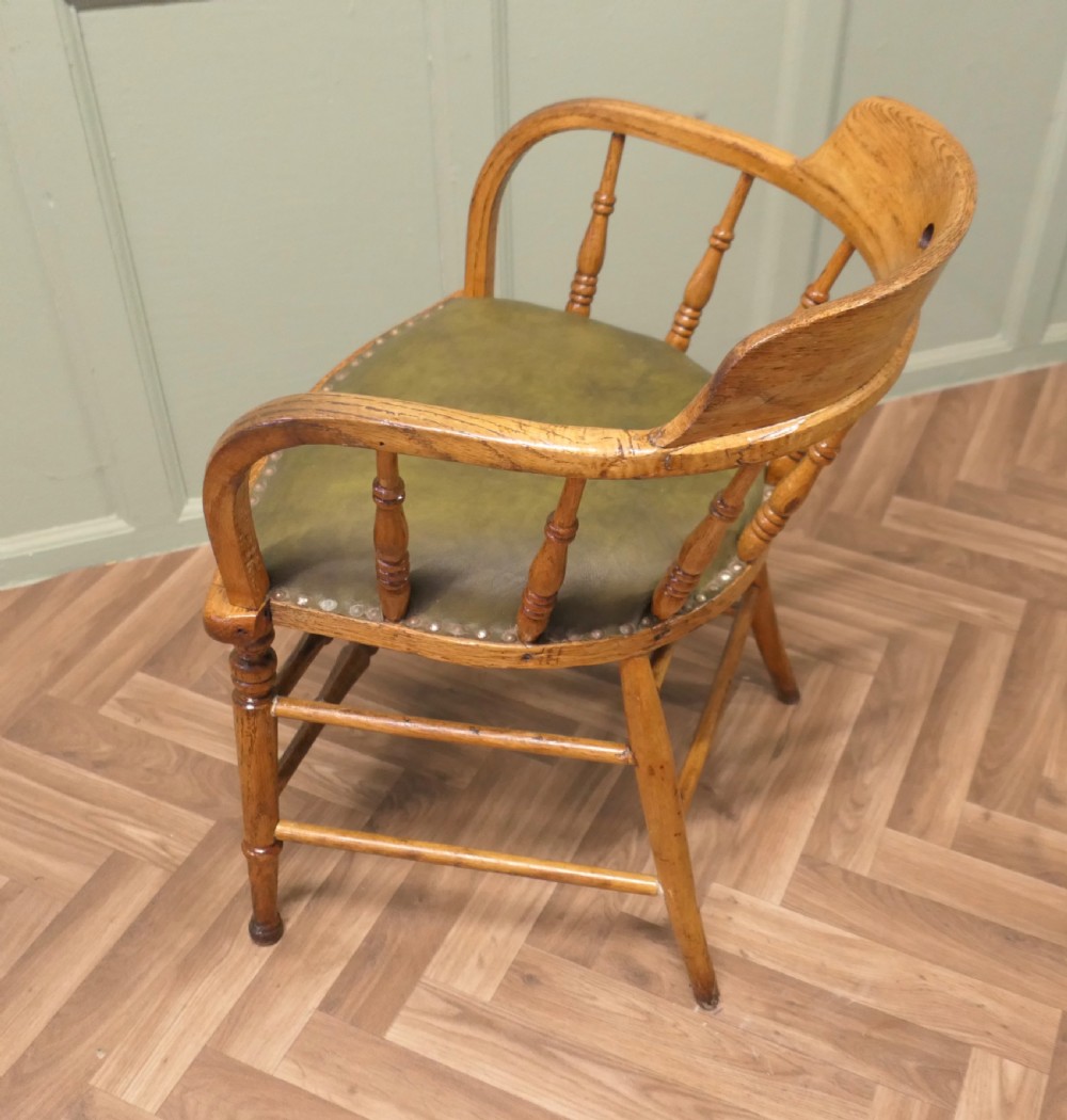 A golden oak smokers bow armchair featuring turned spindles, a steam-bent curved top rail, double stretchers, and a padded green leather seat secured with studs. View VI