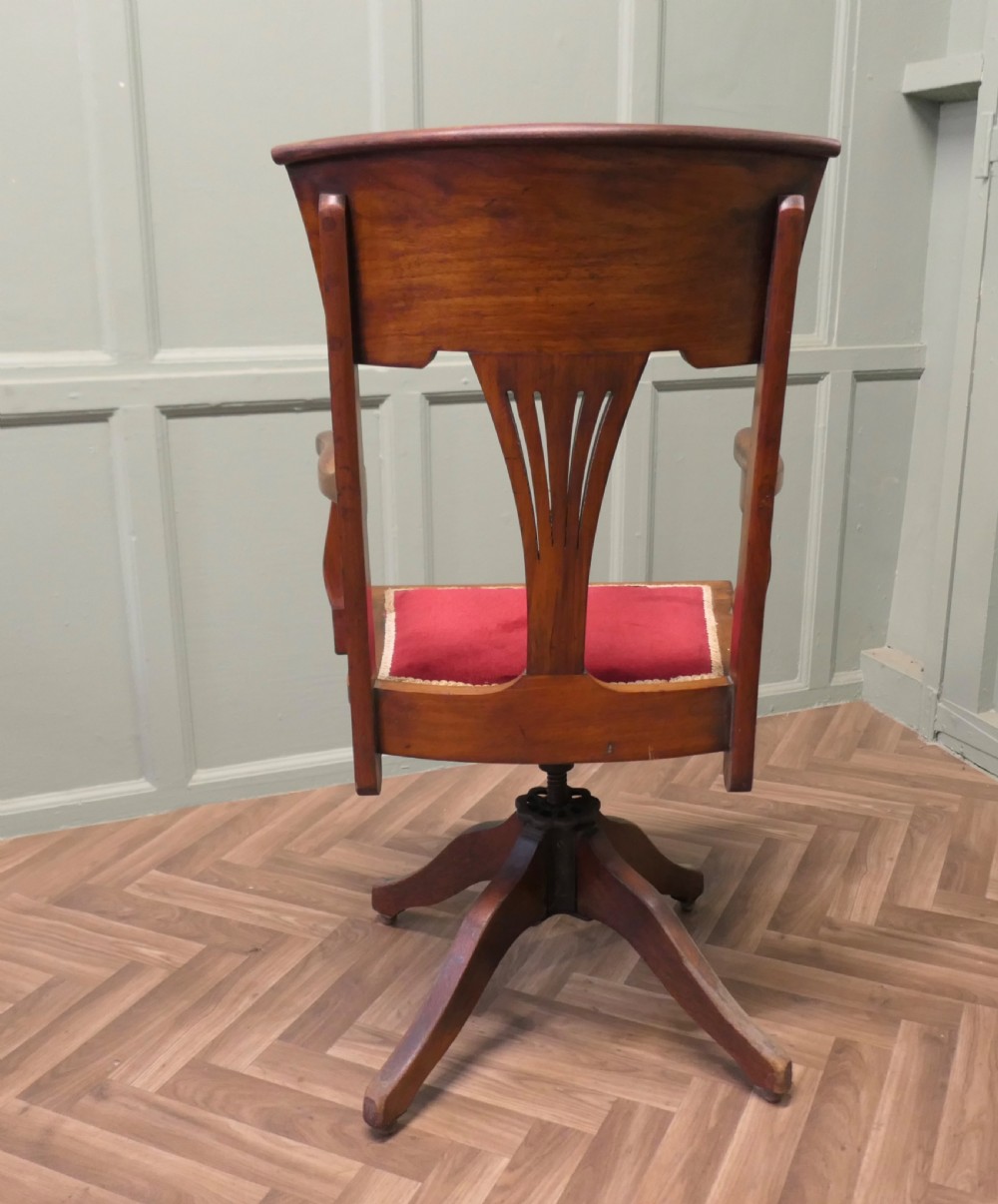 An Edwardian Arts and Crafts walnut revolving desk chair with a curved backrest, carved splat detail, shaped armrests, and a red velvet seat insert. View V